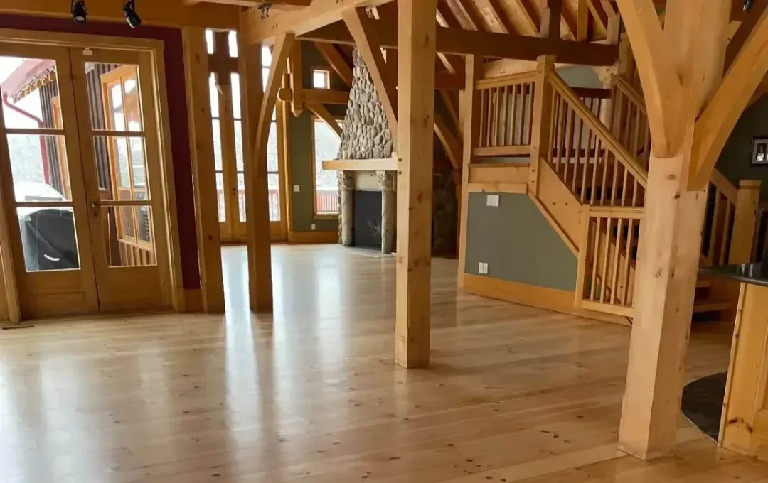 A spacious, rustic home interior featuring wide-plank hardwood flooring with a natural finish, complemented by exposed timber beams, a stone fireplace, and a wooden staircase