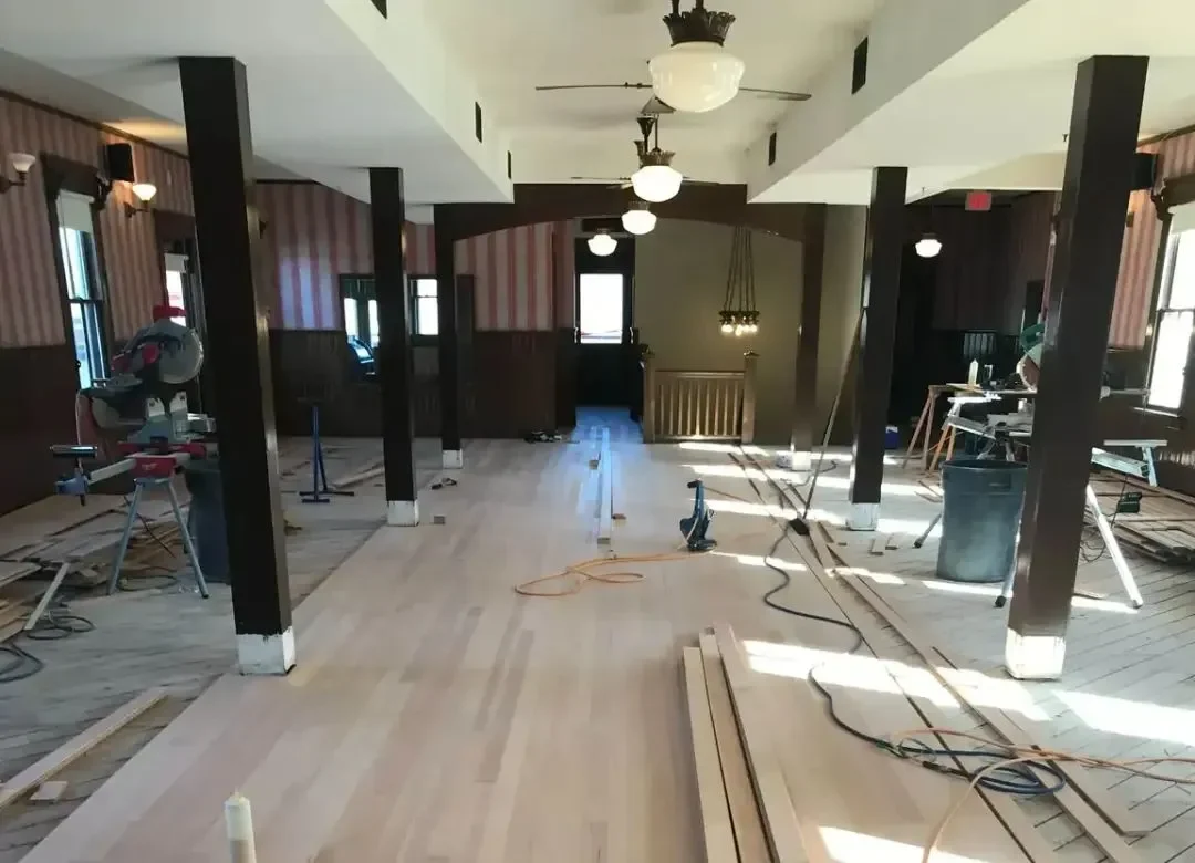 A wide room undergoing hardwood floor installation with light-colored wood planks being laid over a subfloor, surrounded by professional tools like a miter saw and air compressor
